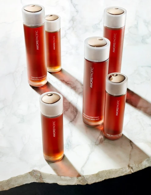 Six amber-colored glass skincare bottles with white and gold caps are arranged on a white marble surface, illuminated by strong side lighting.