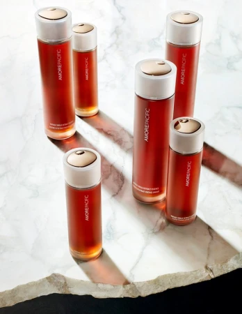 Six amber-colored glass skincare bottles with white and gold caps are arranged on a white marble surface, illuminated by strong side lighting.