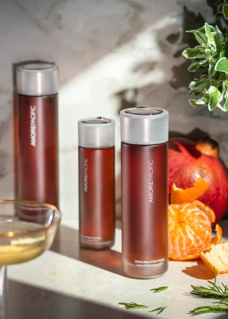 Three Amorepacific skincare bottles are staged with fresh mandarin oranges, pomegranate, and greenery against a light marble backdrop.