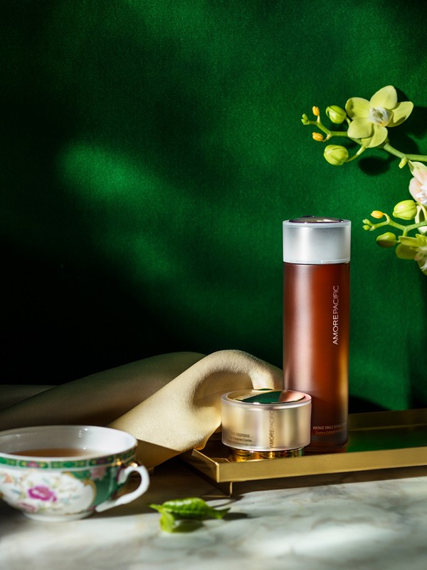 Still life photo of brown glass skincare bottles on a gold tray next to a teacup, green leaves, and flowers against a green background.