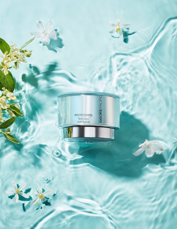 Cosmetic cream jar sits in clear water with ripples and small white flowers and green leaves. Beauty product still life.