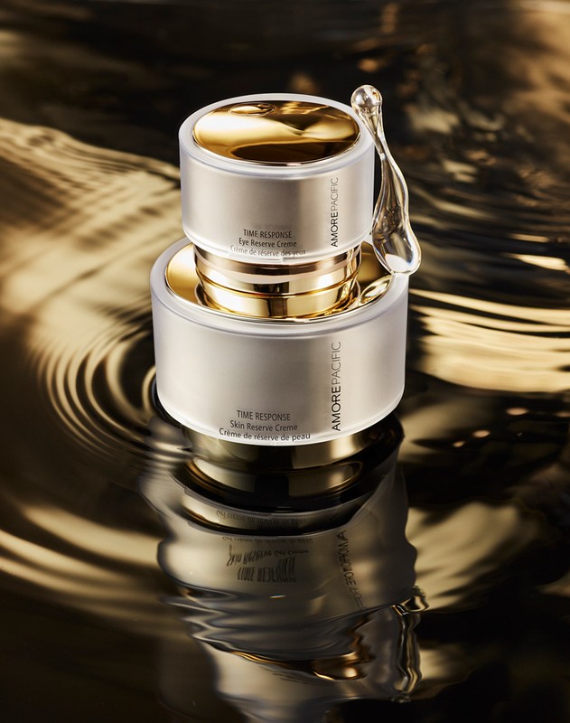 Two AmorePacific skincare cream containers stacked, gold lids, on reflective black and gold liquid surface with rippling effect.
