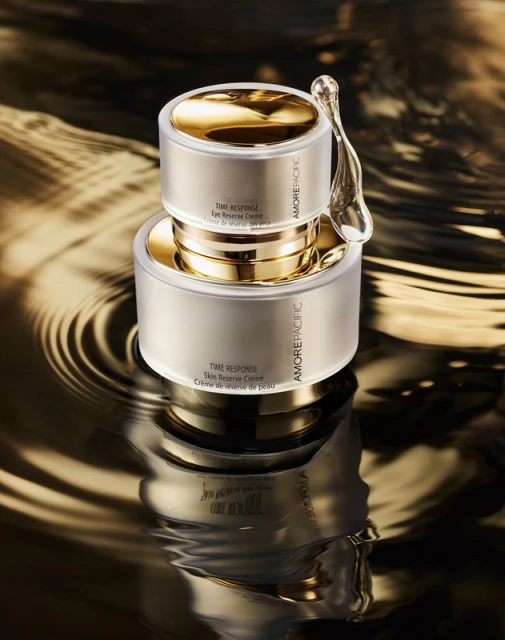 Two AmorePacific skincare cream containers stacked, gold lids, on reflective black and gold liquid surface with rippling effect.