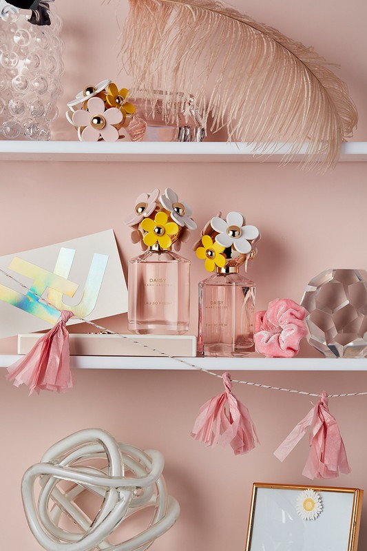 Still life of two Daisy Marc Jacobs perfume bottles with floral caps displayed on a shelf with decorative objects and a garland.
