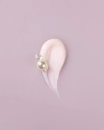 A smear of pink cream cosmetic product on a matching pink background, accented with a white pearl and a clear gemstone.