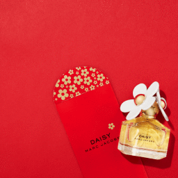 Marc Jacobs Daisy perfume bottle next to a red envelope with gold flower patterns against a vibrant red background.