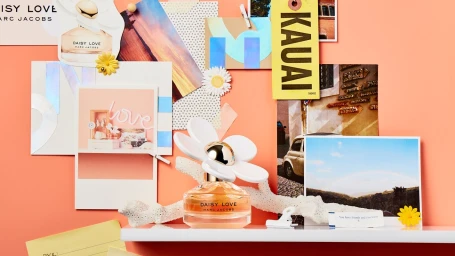Marc Jacobs Daisy Love perfume bottle on white shelf against peach backdrop with collage cards