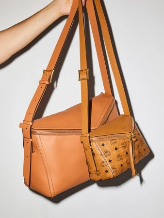 Two leather handbags in tan and brown are displayed, one with the MCM logo pattern, held up by a person's hand against a plain background.