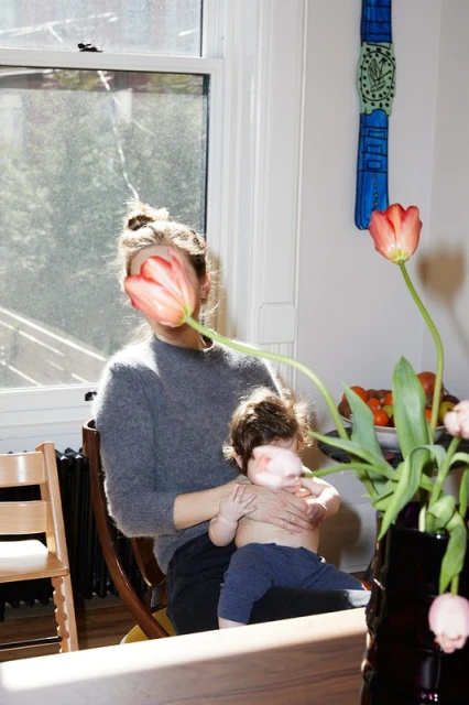 Woman sits in a chair holding a baby, with tulips partially obscuring their faces, indoor setting, bright daylight, cozy atmosphere.