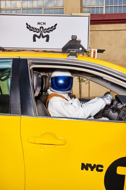 Yellow New York City taxi cab with astronaut driver in reflective white suit beneath MCM billboard in a bright urban scene.