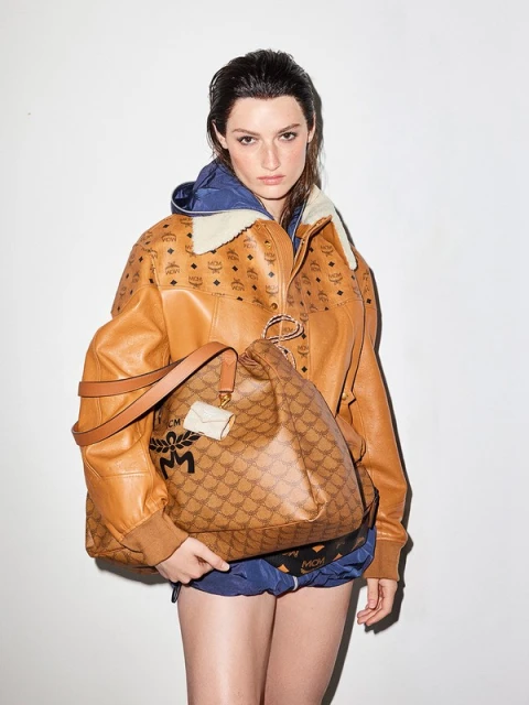 Portrait of a woman wearing a brown printed jacket and shorts, holding a matching handbag in a studio setting with white background.