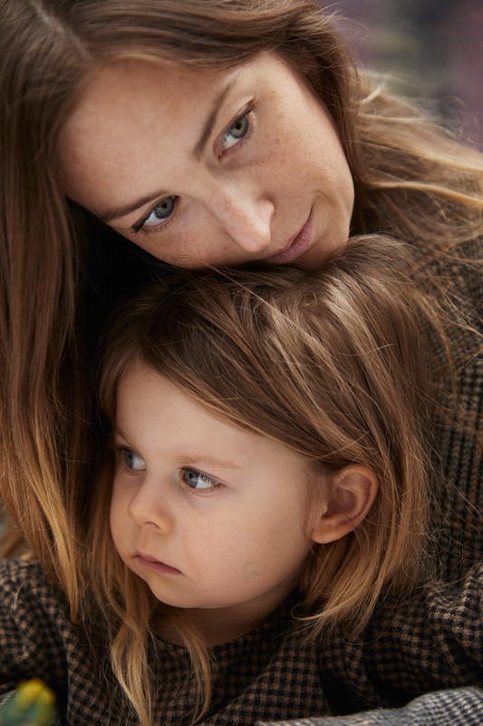 Close-up of a fair-skinned mother and child in an indoor setting. The mother is positioned behind the child, embracing them warmly.
