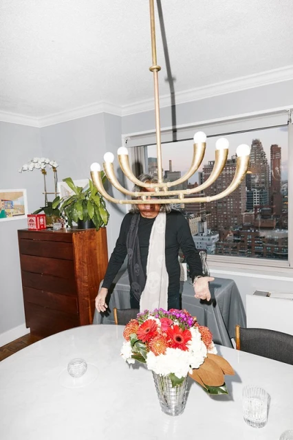 Indoor scene featuring an adult standing near a dining table with a floral centerpiece, chandelier, and a city view through a window.