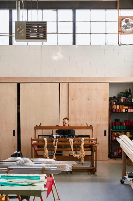 Woman works at loom inside of a large industrial space filled with natural light. Spools of yarn and crafting materials surround her.