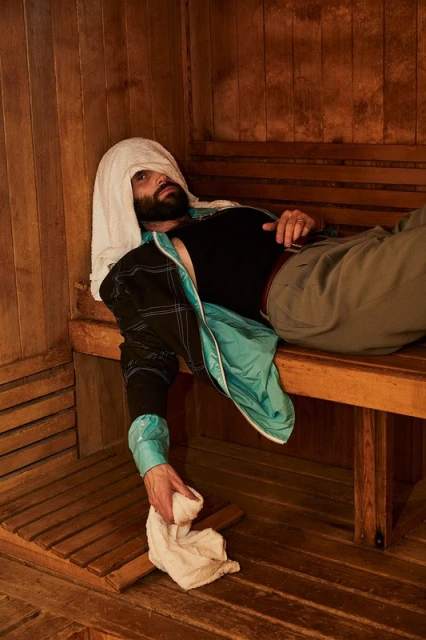 Man in sauna wearing towel on head, relaxing on wooden bench, and holding another towel in hand inside of the wooden sauna.