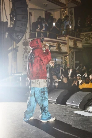 A male music artist on stage performs with a microphone while wearing a red hoodie and patterned pants in front of an audience.