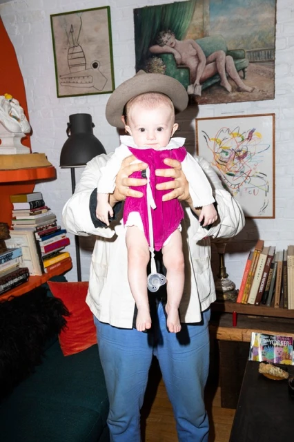 Baby in pink bodysuit and tan hat held by person wearing blue pants and white jacket in room with books and artworks on wall.