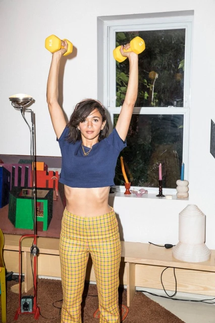 Young woman in workout clothes raises yellow dumbbells overhead in a bright interior. Fitness, exercise, and active lifestyle theme.