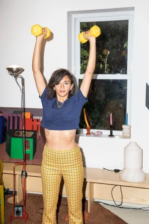 Young woman in workout clothes raises yellow dumbbells overhead in a bright interior. Fitness, exercise, and active lifestyle theme.
