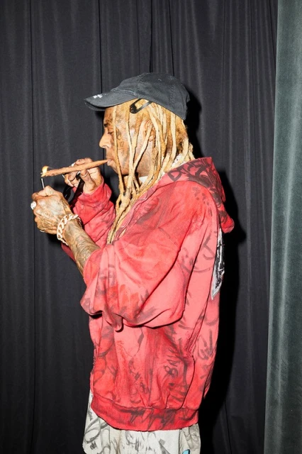 Adult male with blond dreadlocks lights a cigar indoors, wearing a black baseball cap and a red and black patterned hooded sweatshirt.