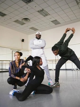 Dance class with four people performing a choreographed routine in a dance studio with mirrors and bright overhead lighting.