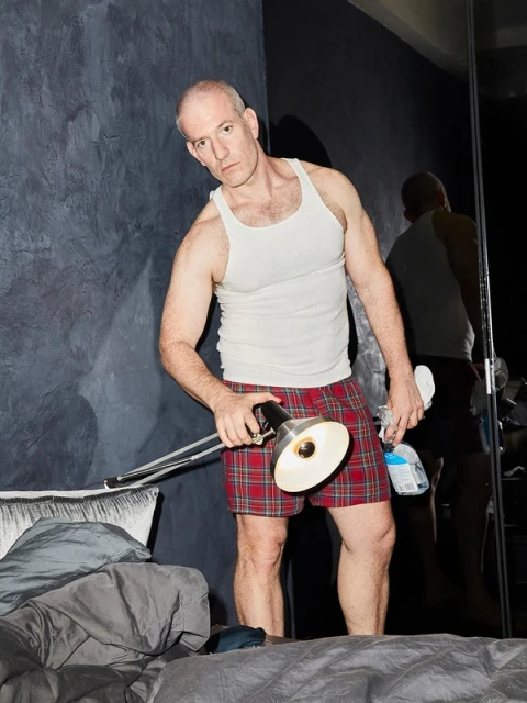 Man in bedroom wearing pajamas, tank top, holding adjustable desk lamp and cleaning supplies near unmade bed and a black textured wall.