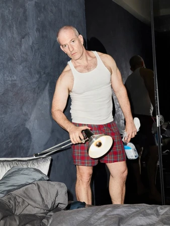 Man in bedroom wearing pajamas, tank top, holding adjustable desk lamp and cleaning supplies near unmade bed and a black textured wall.