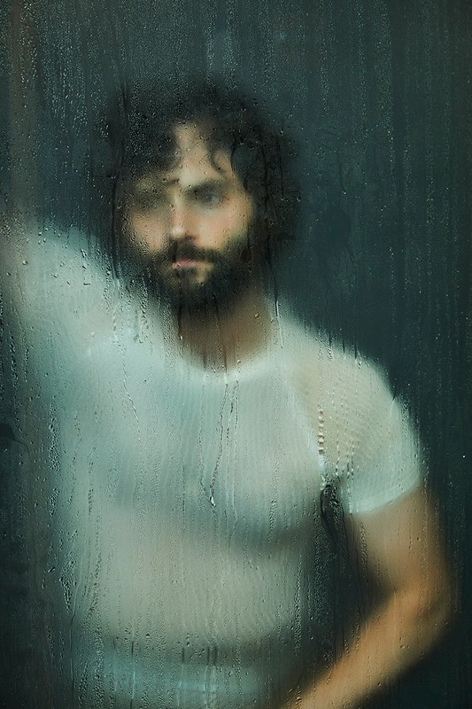 Close-up portrait of a man with dark hair and a beard, wearing a white shirt, standing behind textured glass with water droplets.