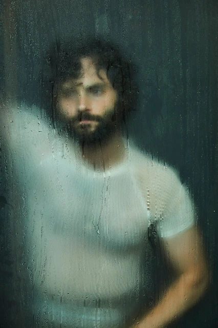 Close-up portrait of a man with dark hair and a beard, wearing a white shirt, standing behind textured glass with water droplets.