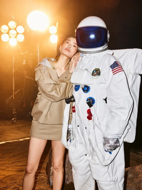 An Asian woman in a tan outfit stands next to a person in a white astronaut suit against a backdrop lit with warm yellow lights.