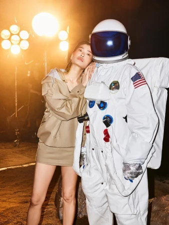 An Asian woman in a tan outfit stands next to a person in a white astronaut suit against a backdrop lit with warm yellow lights.