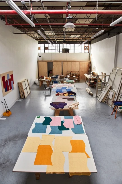 Spacious art studio with tables covered in colorful fabric swatches, ample natural light, and organized art supplies; an inviting creative space.