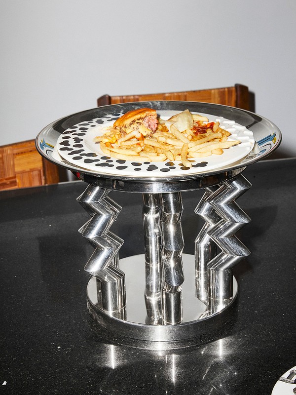Overhead shot of a burger, fries, and garnish on a decorative plate atop a chrome stand, all sitting on a black surface indoors.