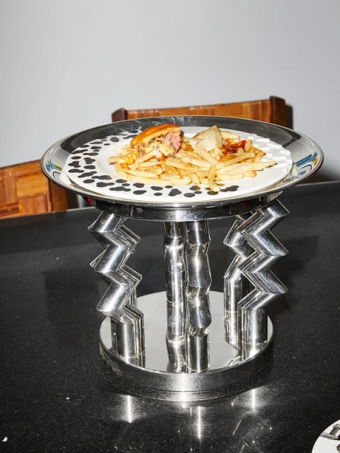 Overhead shot of a burger, fries, and garnish on a decorative plate atop a chrome stand, all sitting on a black surface indoors.