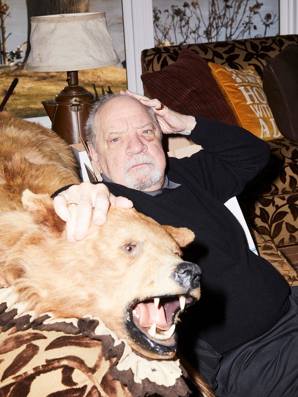 Portrait of elderly man reclining on patterned couch with bear skin rug in an indoor home setting with neutral light.