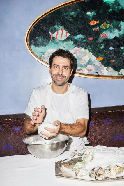 Adult male preparing food at a table with a bowl and tray with oysters against a blue wall with an art piece featuring fish.