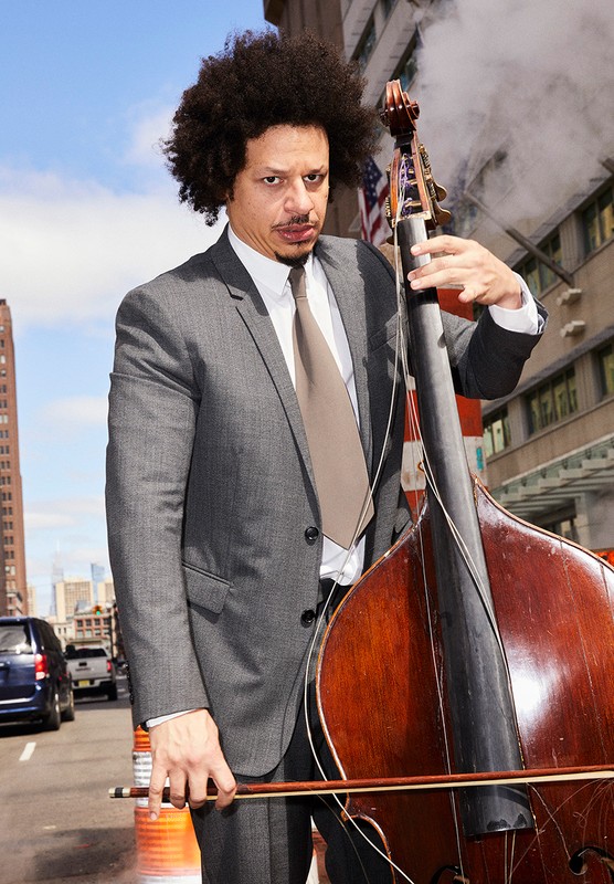 Adult male posing with a cello outdoors on a city street, wearing a suit and tie. Buildings and steam are visible in the background.