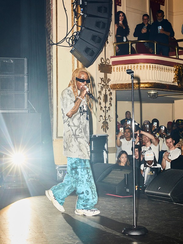 Lil Wayne performing on stage with a microphone, wearing sunglasses and a patterned outfit, with a crowd and stage lighting.