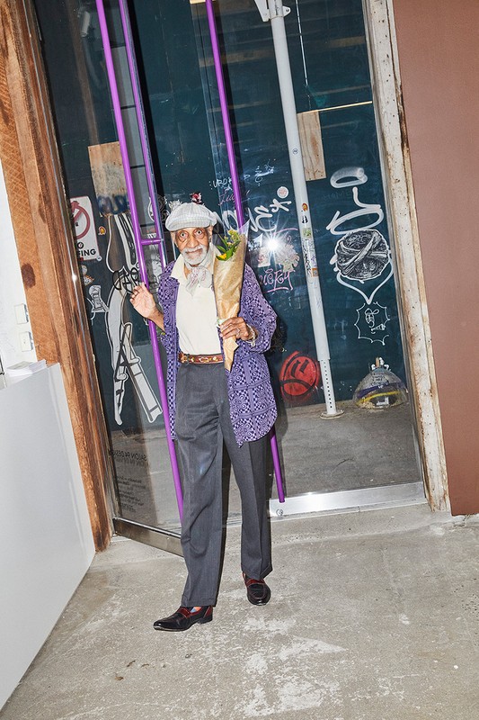 Stylish older man stands outside a door with flowers. He wears a patterned jacket, grey slacks, and hat and gives a slight wave.
