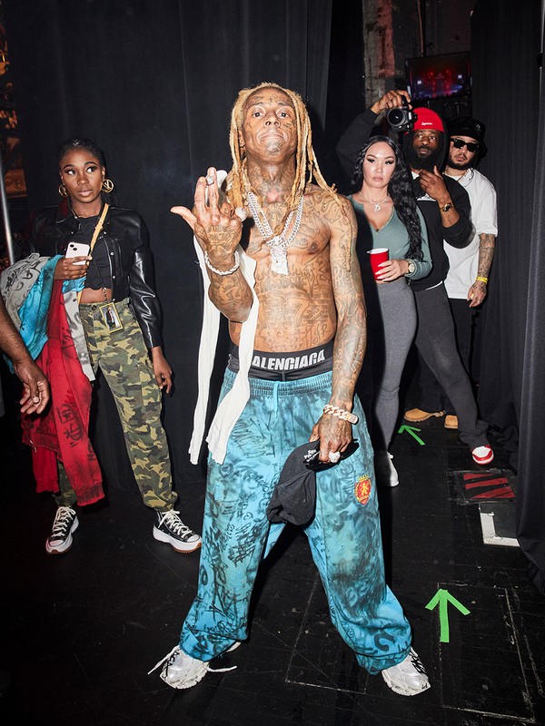 Lil Wayne poses shirtless in patterned pants with jewelry, surrounded by a group in an indoor backstage setting with dark walls.