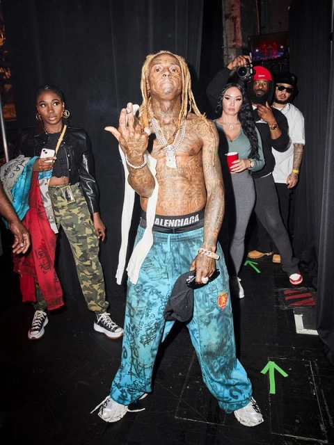 Lil Wayne poses shirtless in patterned pants with jewelry, surrounded by a group in an indoor backstage setting with dark walls.
