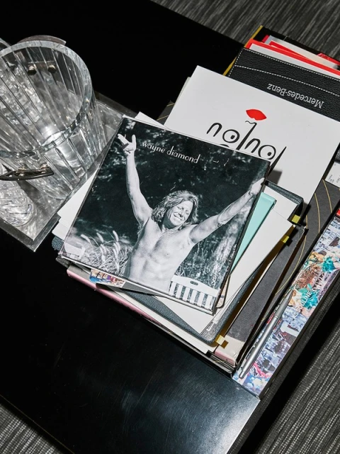 A photograph of various media like a music album of Wayne Diamond, magazines and a container is placed on a black table indoors.