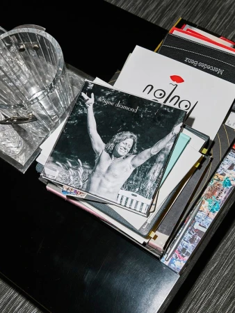 A photograph of various media like a music album of Wayne Diamond, magazines and a container is placed on a black table indoors.
