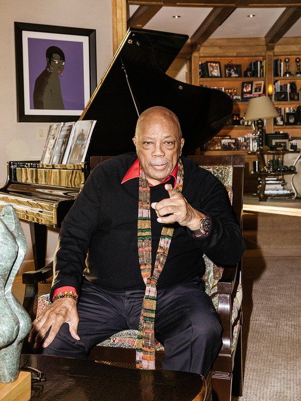 Quincy Jones seated in an armchair pointing forward, wearing a dark sweater and patterned scarf with a grand piano behind him.