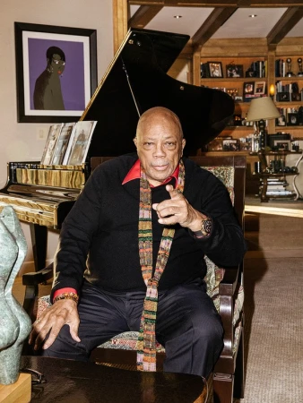 Quincy Jones seated in an armchair pointing forward, wearing a dark sweater and patterned scarf with a grand piano behind him.