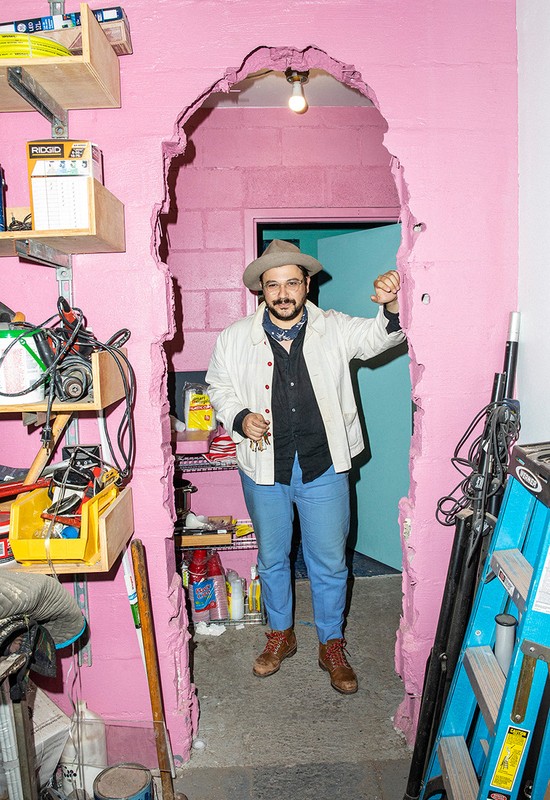 Man in hat stands confidently in a room with a pink cinderblock wall and a doorway, interior design with shelving and tools.