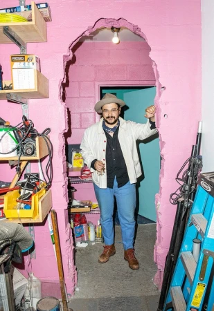 Man in hat stands confidently in a room with a pink cinderblock wall and a doorway, interior design with shelving and tools.