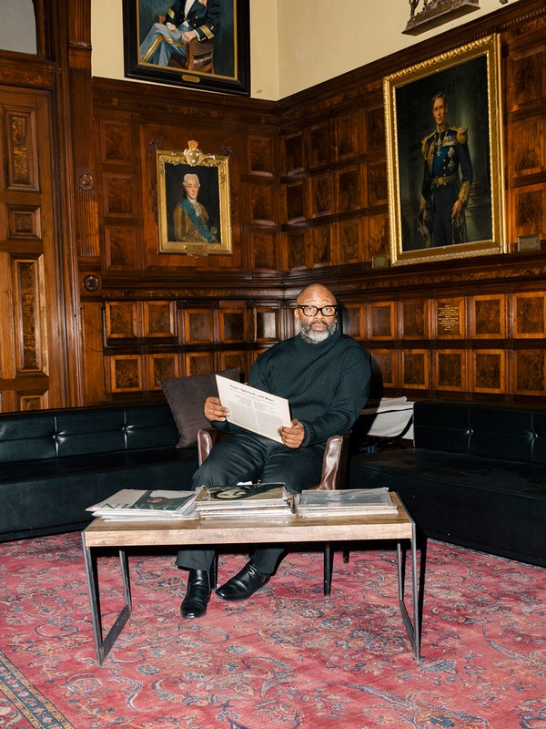 Adult male reading a newspaper indoors in a wood-paneled room with portraits, sitting near a table with vinyl records.