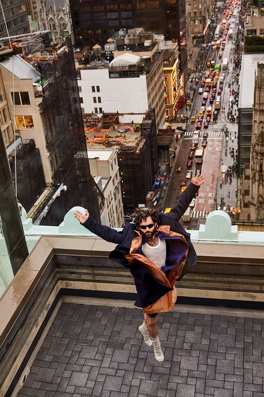 Man jumping on rooftop with urban city background. He is wearing a coat, top, and sneakers. Sunny day with neutral color grading.