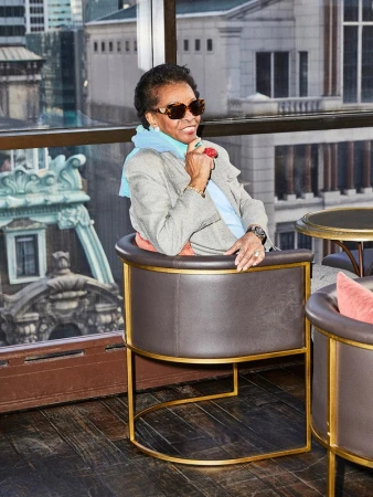 Elderly African-American woman in sunglasses sits in a brown leather chair indoors with a city skyline view from a high-rise window.